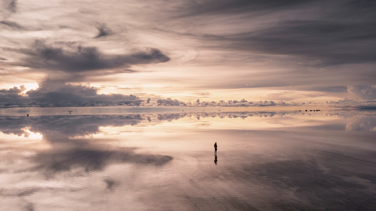 A lone figure stands in a vast, mirrored landscape beneath a dramatic sky, representing the scale of Apollo’s $40T private credit universe—built to fuel growth, enhance resilience, and support retirement income.