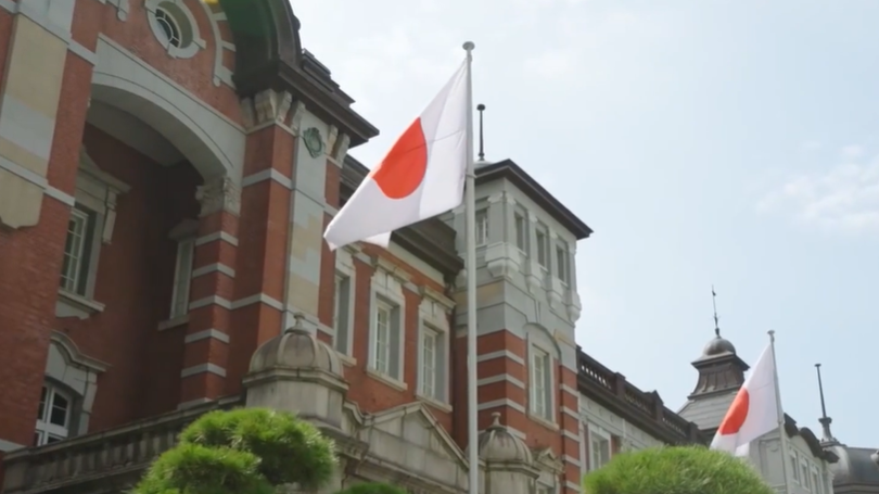 Japan’s Reset Is Rational, Not Cultural Japanese flags displayed outside a historic red-brick building with modern office towers in the background, representing Tokyo’s financial district and global capital markets activity in an urban setting.