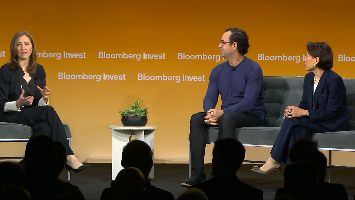 Private Credit, Now in an ETF Panel discussion at Bloomberg Invest featuring three speakers seated on stage in front of a orange backdrop with the Bloomberg logo discussing private credit and retail access to private markets, with audience silhouettes in the foreground.