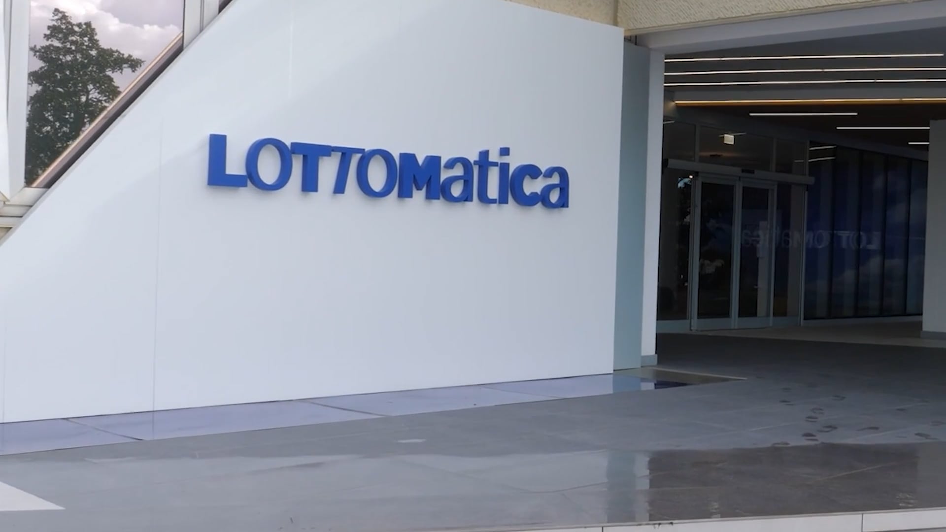 Lottomatica: Transforming a Leader in Italian Gaming Exterior view of Lottomatica's headquarters entrance, featuring the company’s blue logo on a white wall and glass doors leading into the building.